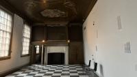 Een historische kamer met een hoog gedecoreerd plafond en grote ramen. Met een zwart-wit geblokte vloer, een grote open haard met sierlijke details en twee moderne stoelen tegen een eenvoudige muur. De kamer wordt zacht verlicht door natuurlijk licht.
