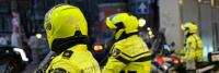 Drie politieagenten in felgele uniformen en helmen zitten op motoren. Ze staan geparkeerd op een stadsstraat, mogelijk houden ze het gebied in de gaten. De reflecterende jassen tonen "Politie” en een embleem op de mouwen.