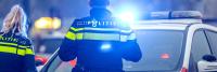 Twee politieagenten in uniform staan bij een politieauto met zwaailichten. De scène lijkt zich in een stedelijke omgeving af te spelen en de agenten kijken weg van de camera. Op de politieauto staat "Politie".