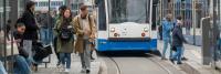 Mensen staan en lopen op een tramplatform in een stedelijke omgeving. Een tram staat stil en sommige mensen stappen in terwijl anderen voorbij lopen. De scène bestaat uit mensen die op banken zitten en anderen die met hun telefoon of met elkaar communicer