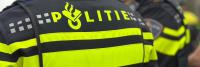 Een close-up van de achterkant van een politie-uniform met het woord "POLITIE" in vette gele letters. Het jasje heeft reflecterende gele en zwarte strepen. Een andere agent staat aan de zijkant, op dezelfde manier gekleed.