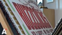 Close-up van een stapel posters met gestileerde tekst. De voorgrond richt zich op rode en witte letters. Een hoek van een kamer met een houten plank en een fotolijst is zichtbaar op de achtergrond. Een logo met de letter "A" staat in de linkerbenedenhoek.