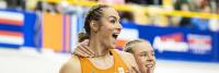 Twee atleten in oranje uniformen vieren met vreugdevolle uitdrukkingen na een sportevenement. De een heeft gevlochten haar en lacht breed, terwijl de ander hen omhelst en ook lacht. Een wazige menigte is zichtbaar op de achtergrond.