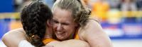 Twee vrouwen in oranje sportkleding omhelzen elkaar en lachen vrolijk. Het gezicht van een vrouw is zichtbaar en haar haar is naar achteren getrokken. Ze zijn binnen, met een wazige achtergrond met mensen en structuren.
