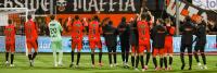 Een voetbalteam in oranje en zwarte uniformen klapt en juicht terwijl ze tegenover een menigte staan. Ze staan op een veld met een doelpaal aan de linkerkant. Een spandoek met de woorden "Riverside Maffia" hangt boven hen.