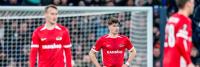 Drie voetballers in rode shirts staan op een veld voor een doel. De centrale speler heeft zijn handen op zijn heupen. De shirts tonen een logo en de tekst "KANSINO." De achtergrond toont een publiek en wazige omgevingsdetails.