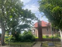 Een klein bakstenen monument met een metalen kruis staat voor een huis met een rood pannendak, omringd door bomen en groen onder een gedeeltelijk bewolkte hemel. Links is een straatnaambord zichtbaar.