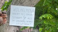 Aan een boom hangt een handgeschreven bord in het Nederlands. Op het bord staat: "Wilhelmina heeft mij geplant. Schaam je als ik vandaag word afgedankt." Op de achtergrond zijn groene bladeren en boomschors te zien.