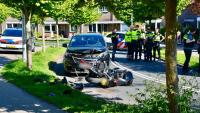 Een beschadigde zwarte auto en een vernielde motorfiets staan in een woonstraat. Verschillende politieagenten staan in de buurt en aan de linkerkant staat een politieauto geparkeerd. Puin ligt verspreid over de weg.