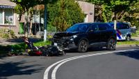 Een zwarte SUV met een beschadigde voorkant staat stil op de weg na een botsing met een motorfiets die op zijn kant ligt. Een politievoertuig staat vlakbij geparkeerd en op de achtergrond zijn woonhuizen zichtbaar.