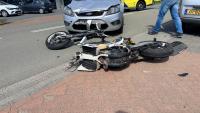 Een beschadigde motorfiets ligt op zijn kant bij een stoeprand na een botsing met een zilverkleurige auto, die een kapotte voorbumper heeft. Er liggen brokstukken op de weg en vlakbij loopt iemand in spijkerbroek.