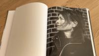 Een open boek op een houten oppervlak toont een zwart-witfoto van een vrouw in profiel, kijkend naar rechts, met een bakstenen muur op de achtergrond. De linkerpagina van het boek is blanco.
