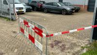 Een metalen barrière met rood en wit lint blokkeert een parkeerplaats met meerdere geparkeerde auto's. Op de grond zijn gele bewijsmarkeringen en een donkere vlek zichtbaar.