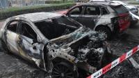 Op een parkeerplaats staat een zwaar uitgebrande auto naast een beschadigde rode SUV. De grond is verschroeid en het gebied is afgezet met politielint waarop „NIET BETREDEN“ staat. Op de achtergrond zijn struiken en een trottoir te zien.