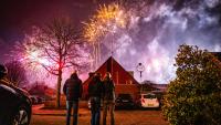 ’s Nachts staan drie mensen op een parkeerplaats en kijken naar kleurrijk vuurwerk in de lucht boven een huis. Er zijn kale bomen, geparkeerde auto’s en een struik te zien, terwijl de achtergrond gevuld is met rook van het vuurwerk.